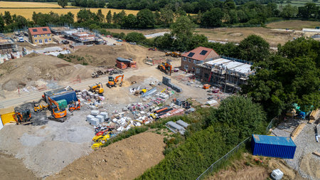 Aerial view of a residential construction site with multiple houses under various stages of development. Excavators, building materials, and shipping containers are visible.の写真素材