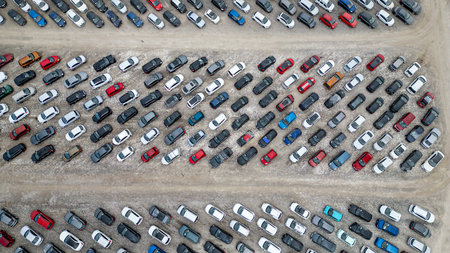 An overhead aerial view shows a large outdoor parking lot filled with numerous vehicles. The cars...