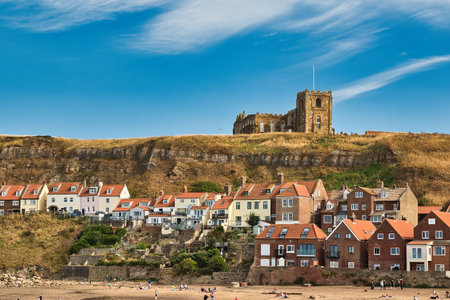 Coastal town with buildings nestled on a hillside beneath a prominent church structure. A sandy beach with scattered people lies in the foreground  in Whitby, UK.の写真素材