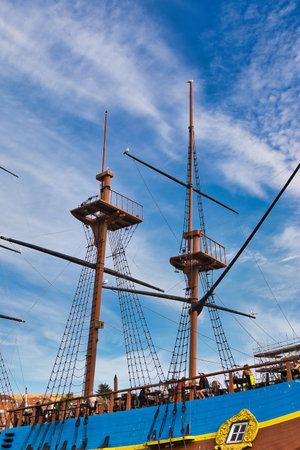 A close-up, low-angle view of a wooden ship's masts and rigging against a blue sky with wispy...