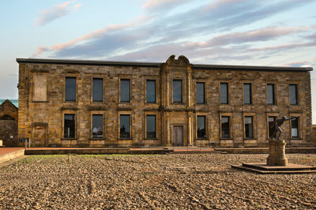 A weathered stone building with a central pediment and numerous windows stands on a pebbled courtyard. A bronze statue is placed in front.の写真素材
