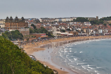 A wide, sandy beach is dotted with people and leads to a blue ocean with breaking waves. Buildings line the shore, with a Ferris wheel visible  in Scarborough, UK.の写真素材