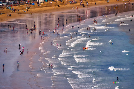 A high-angle shot of a crowded beach with people on the sand, in shallow water, and in the ocean, with waves and reflections  in Scarborough, UK.の写真素材