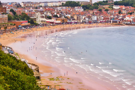 A coastal town with a sandy beach dotted with people, buildings, and a Ferris wheel, against a pale sky  in Scarborough, UK.の写真素材