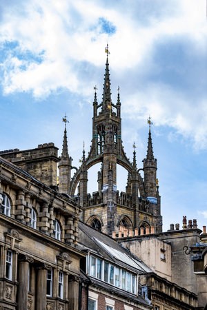 Gothic architecture dominates the frame with a detailed stone church spire and ornate building facades against a blue sky with white clouds  in Newcastle upon Tyne, UK.の写真素材