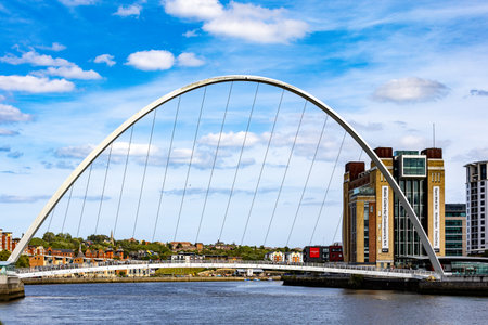 A white arched footbridge spans a river, with a cityscape and modern buildings on either side. Bright blue sky with scattered clouds  in Newcastle upon Tyne, UK.の写真素材