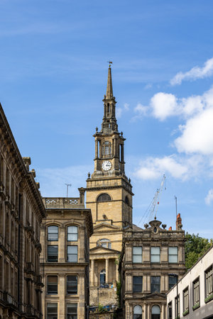 A tall, ornate clock tower with a spire dominates the center of the frame, surrounded by historic stone buildings under a bright blue sky with scattered clouds  in Newcastle upon Tyne, UK.の写真素材