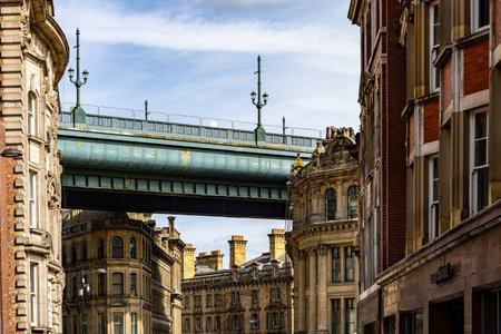 A teal bridge spans across a street lined with ornate, old buildings under a partly cloudy sky in Newcastle upon Tyne, UK.の写真素材