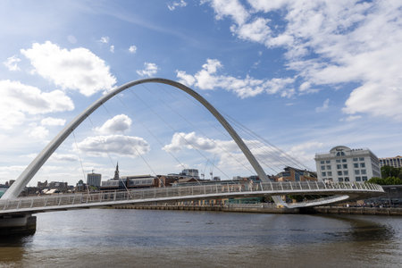 A modern white arch bridge spans a river with a cityscape and cloudy blue sky  in Newcastle upon Tyne, UK.の写真素材
