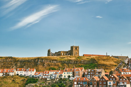 A vast blue sky with wispy clouds above a grassy hill topped with ruins of a church. Below, a village of houses with red roofs is nestled on the hillside  in Whitby, UK.の写真素材