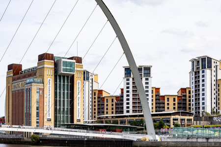 Modern buildings, a cable-stayed bridge, and a river are visible under a bright, cloudy sky in Newcastle upon Tyne, UK.の写真素材