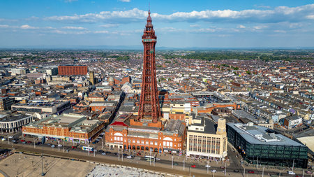 An aerial view of a bustling coastal city, dominated by a tall, red iron tower. Buildings in various architectural styles and colors fill the landscape under a bright blue, cloudy sky.の写真素材