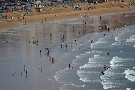 An aerial view of a busy beach with people walking, playing, and swimming in the ocean. The wet sand reflects the sky, and waves crash on the shore  in Scarborough, UK.の写真素材