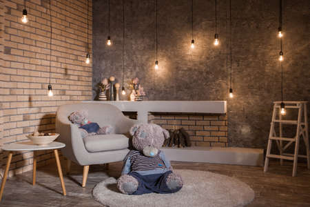 Loft style room with burning bulbs garland, yellow brick wall, gray armchair and modern fireplace. Big teddy bear sitting on the floor. Horizontalの写真素材