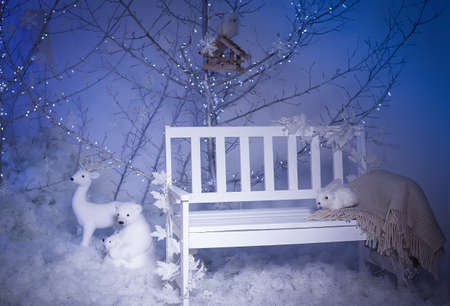 Winter decoration. Silver tree decorated with snowflakes and Christmas lights. White bench with frozen leaves and sled with teddy bear on snowの写真素材