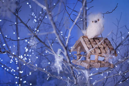 White owl sitting on the birdhouse among silver tree branches decorated with Christmas lights, snowflakes. Blue Background. New Year scene.の写真素材