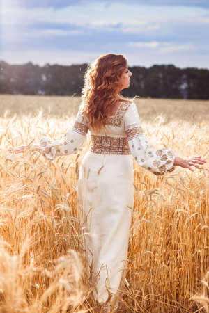 Pretty woman wearin? a embroidered dress with braid standing with her back to the camera among of golden field and pushes wheatの写真素材