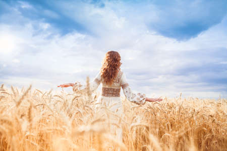 Pretty woman wearin? a embroidered dress with braid standing with her back to the camera in the field and pushes wheatの写真素材