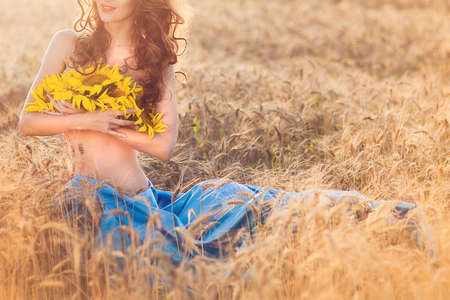 Unrecognized young girl pressed to itself sunflowers sitting at the wheat field. Nature concept. Horizontalの写真素材