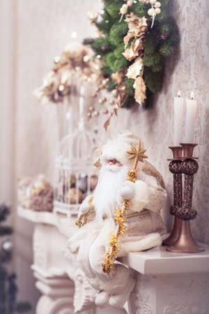 White Santa sitting on classical fireplace with golden stick under Christmas wreath. Xmas background. Winter concept.の写真素材