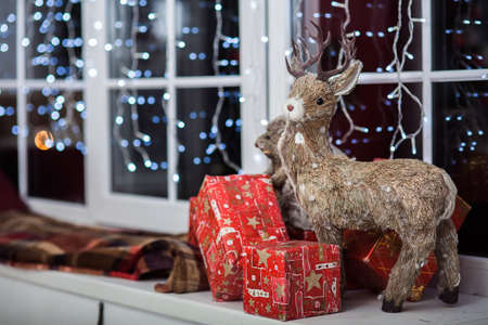 Decorative deer standing on a windowsill near red gift boxes. Lightning garland at the backgroundの写真素材