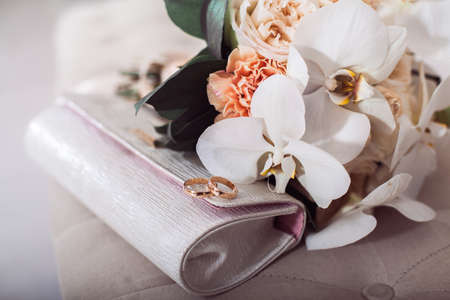 two golden wedding rings on bridal clutch near white flowers bouquet.の写真素材