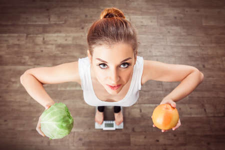 Caucasian smiling girl stands on scales and holding a cabbage and grapefruit in her raised hands. Healthy lifestyle. Vegan and raw food diet concept. View from above.の写真素材