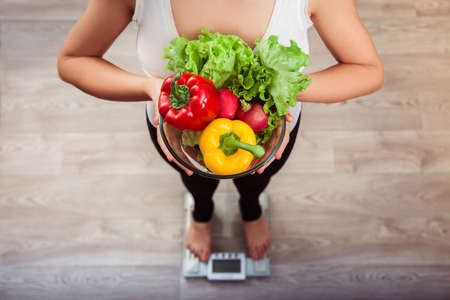 unrecognizable slim girl checking weight on scale holding glass bowl with sweet pepper, fresh...