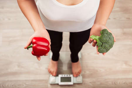 unrecognizable slim woman checking weight on scale and holding vegetables. Weight loss and diet concept. Vegan girl counts caloriesの写真素材
