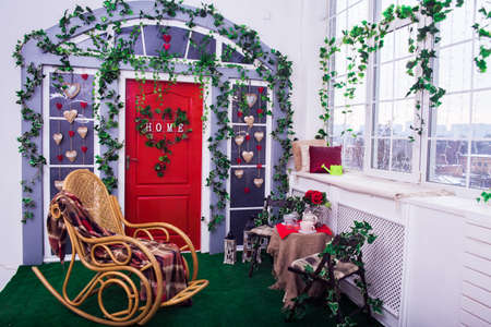 Porch with red door and climbing ivy on the wall. Wicker rocking chair at the spring terrace. Wide angleの写真素材
