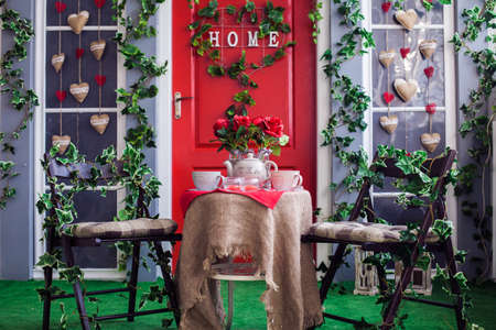 two chairs and a table served for tea ceremony in front of red door entwined with green ivy. Romantic tea partyの写真素材