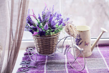 Small decorative bicycle with wicker basket pour of spring lavender bouquet and white watering can on the windowsill covered lilac plaid. Close-upの写真素材