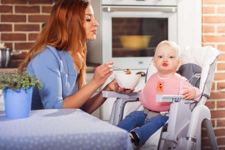 Little baby girl sits in high chair and throwing a kiss while beautiful mother feeding her with spoon. Family and motherhood concept. Horizontalの写真素材