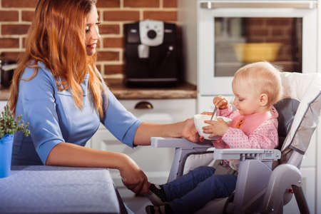 Little baby girl sits in high chair and makes the first attempt to eat with a spoon. Her beautiful mother is happy. Childhood and motherhood concept. Horizontalの写真素材