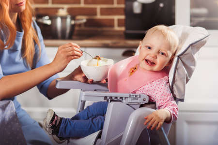 Little baby girl sits in high chair and won't  to eat the food and cry. Mother feeding her with spoon. Family and motherhood concept. Horizontalの写真素材