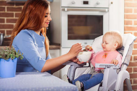Beautiful mother in blue dress feeding with spoon her cute baby girl witch sits in high chair. Family and motherhood concept.  Close up. Horizontalの写真素材