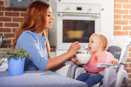 Beautiful mother in blue dress feeding with spoon her cute baby girl witch sits in high chair. Family and motherhood concept.  Close up. Horizontalの写真素材