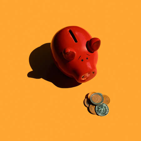 Sad piggy bank looks at euro coins. Red piggy bank with bunch of coins. Minimalist pig on Orange background.の写真素材