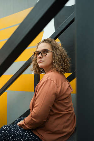 Young adult millennial with curly hair sits on steps. Student looks away. Lifestyle. The girl is...