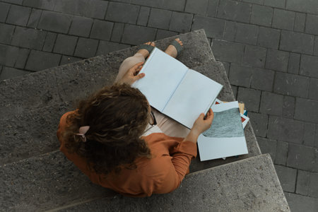 Young adult millennial with curly hair sits on steps. Student reads book. Lifestyle in city