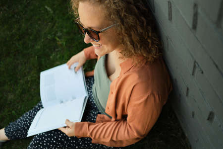 Young adult millennial with curly hair smiles and sits on grass. Student reads book. Lifestyle. The...
