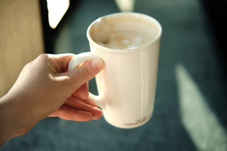 Hand holds cup of coffee, sunlight from window. Woman takes break from remote work to rest and drink coffee.の写真素材