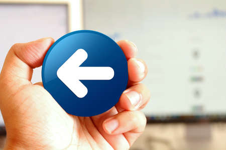 Back arrow icon blue round button holding by hand infront of workspace background closeup business conceptの写真素材
