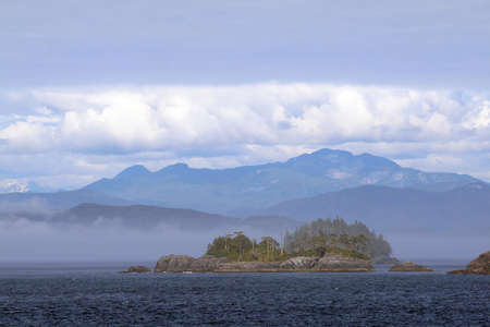 Rocky Islands in Alaskas Inside Passage on a sunny, hazy afternoon with mountains and clouds in the background.の写真素材