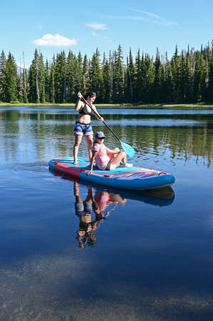 Two young women on standup paddle board on Scott Lake, Oregon with Middle and North Sisters...