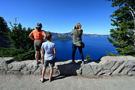 Three women enjoy and photograph view of Crater Lake in Crater Lake National Park, Oregon on a clear...