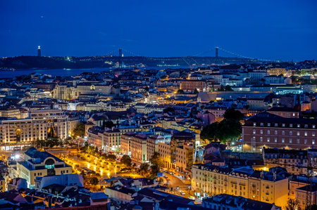 Illuminated buildings of Lisbon, Portugal in deep afternoon twilight with Tagus River and bridge in background from Miradouro da Senhora do Monte.の写真素材