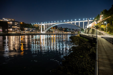 Brightly illuminated Arrabida Bridge and surrounding buildings reflected in calm water of Douro River in Porto, Portugal at night.の写真素材