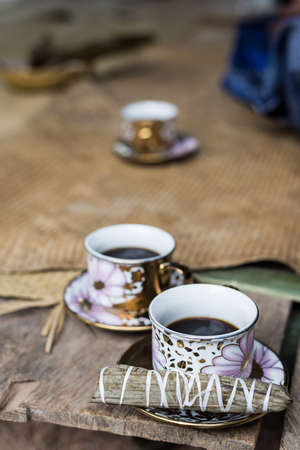 Selective focus on one of three cups of black coffee with traditional snack (sweet rice rolled inside palm leaf) on wooden bench. Sulawesi, Indonesia.の写真素材