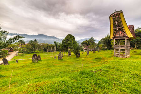 Tipical boat shaped roofs of an ancient traditional wooden building around a cluster of megaliths on the hill of Rantepao, Tana Toraja, South Sulawesi, Indonesia. Wide angle shot, awesome cloudy sky.のeditorial素材